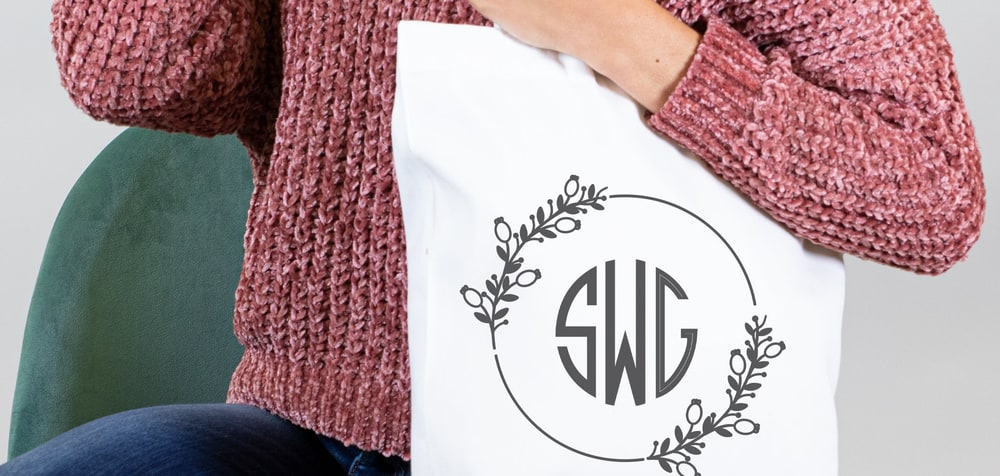 A woman in a pink knit sweater holding a white canvas tote printed with a three-letter circle monogram