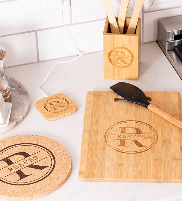A bamboo cutting board, cork trivet, coaster and utensil holder engraved with a family monogram on a kitchen counter