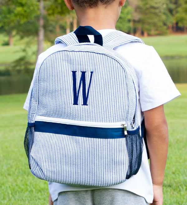 A child outdoors wearing a blue and white seersucker backpack monogrammed with the letter W