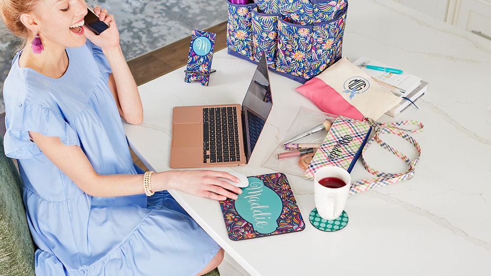 A woman on the phone at a white marble desk with a monogrammed mouse pad, phone stand, pouch and a printed organizer tote