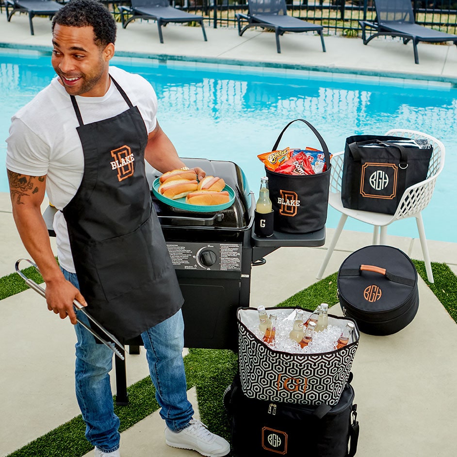 A man grilling hot dogs beside a pool, surrounded by monogrammed black cooler totes, a drinks tub and a personalized apron