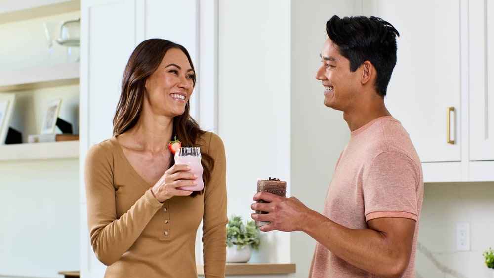 A smiling woman and man chat in a bright white kitchen, she holding a strawberry shake and he a chocolate shake