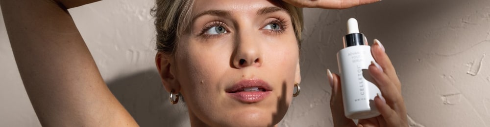 A woman with her arm raised above her head holds a white bottle of Celletoi Advanced Youth Serum beside her face