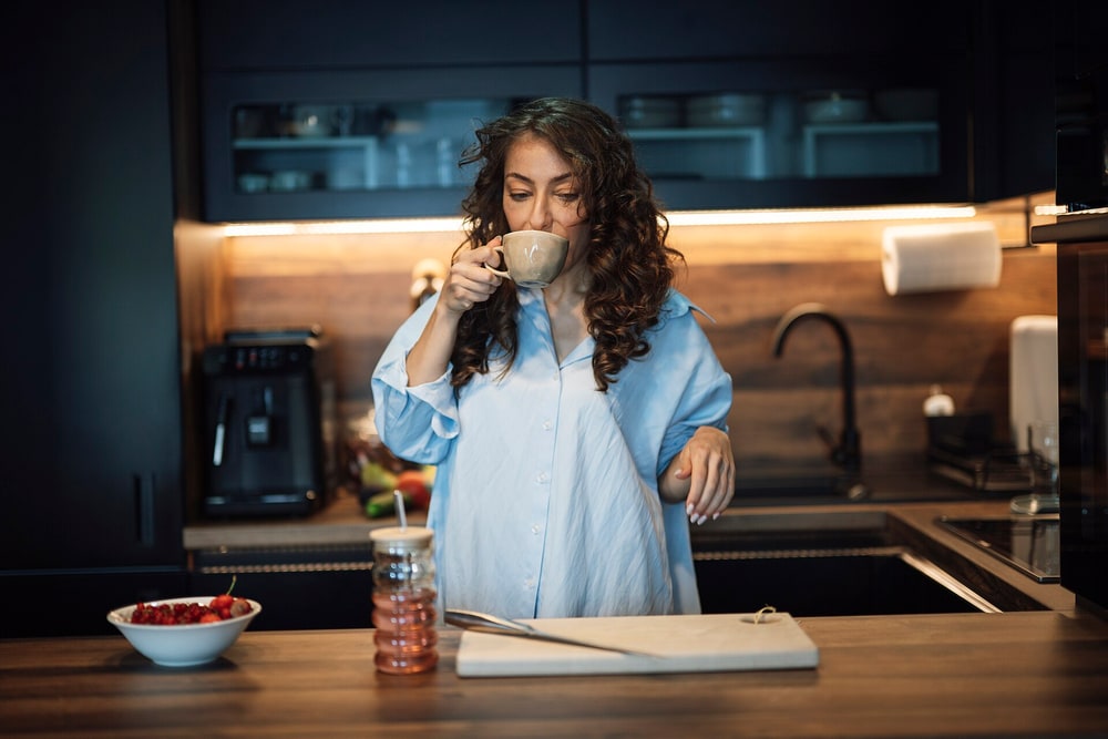 Woman in a blue shirt sipping a cup of coffee at a dark wood kitchen counter with a bowl of berries