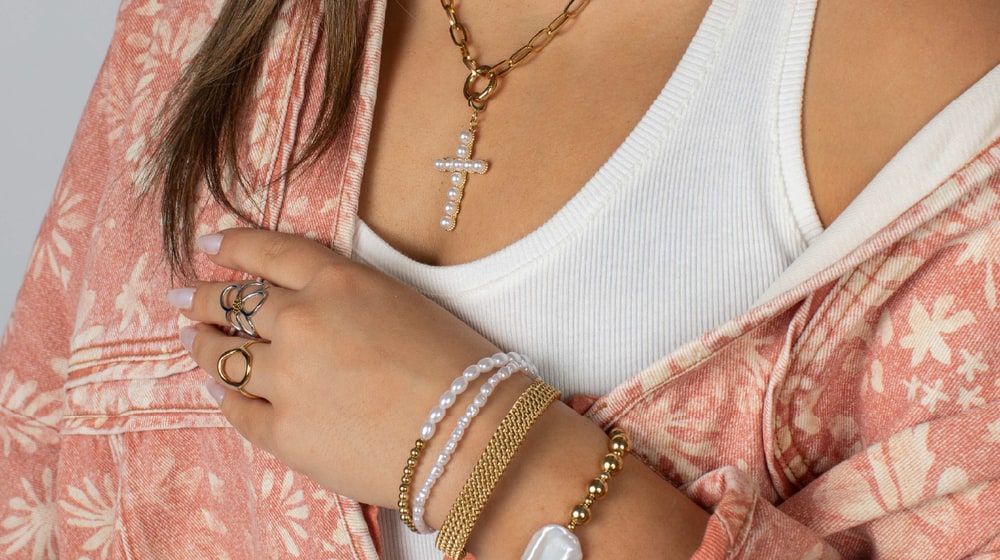 Woman in a pink floral jacket wearing a gold paperclip chain with a pearl cross pendant, layered pearl and gold bracelets and stacked rings