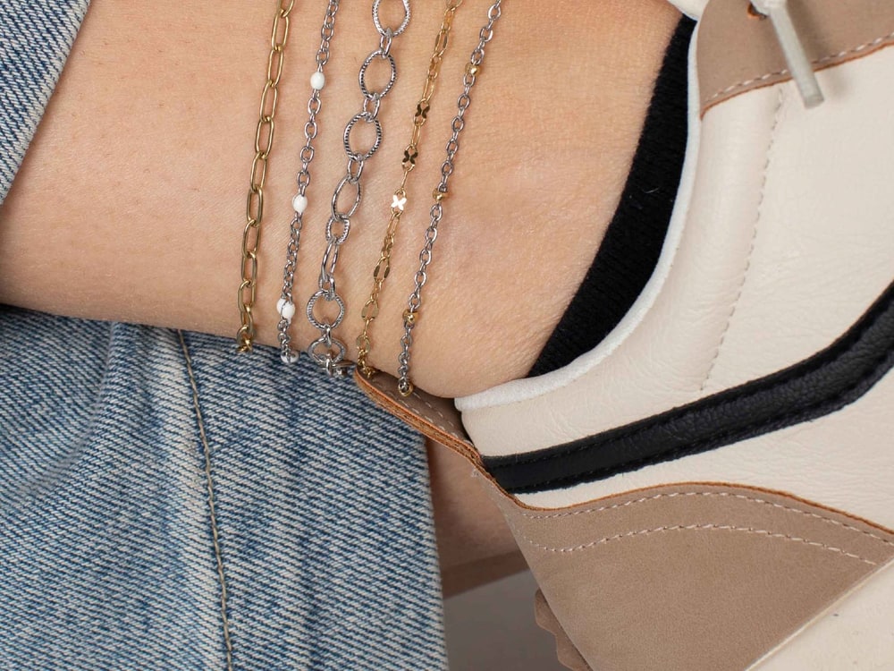 Five fine gold and silver chain anklets stacked above a cream sneaker and rolled denim