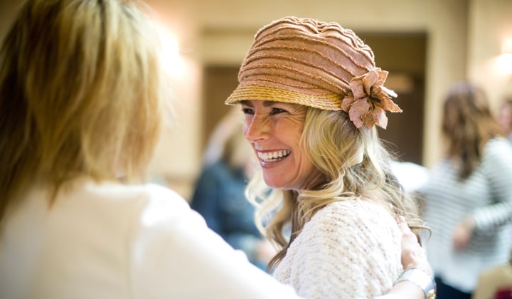 A smiling woman in a knitted cloche hat greets a friend at a busy Jewel Kade Stylist gathering