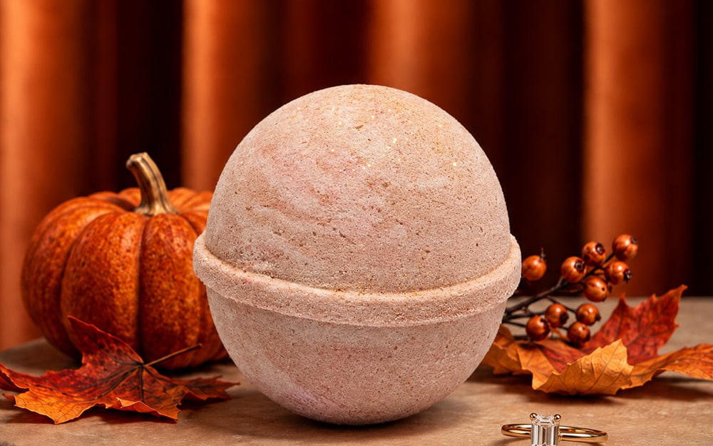 A pale pink JewelScent bath bomb on a stone table beside a gold emerald-cut ring, a small pumpkin and autumn leaves