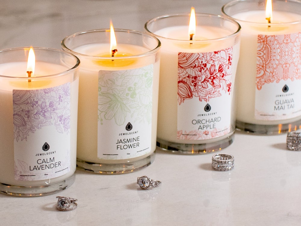 Four lit JewelScent 18 oz candles in Calm Lavender, Jasmine Flower, Orchard Apple and Guava Mai Tai with silver rings on the marble beside them