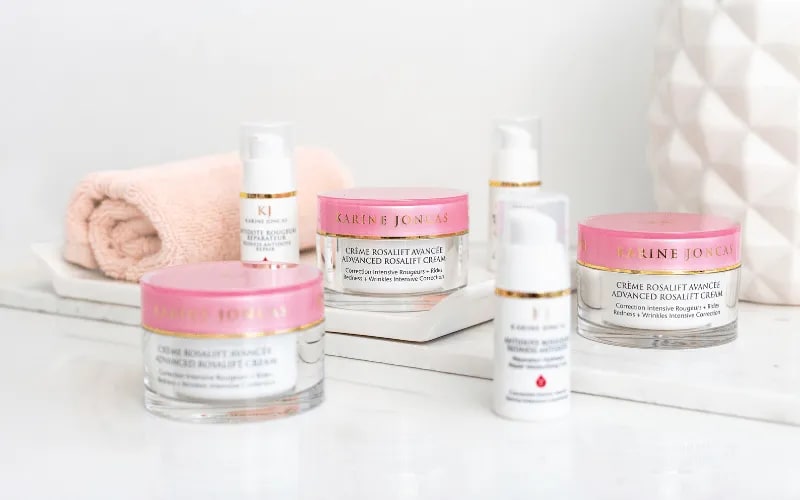 Karine Joncas Advanced Rosalift creams and serums with pink lids on a white marble shelf beside a rolled towel