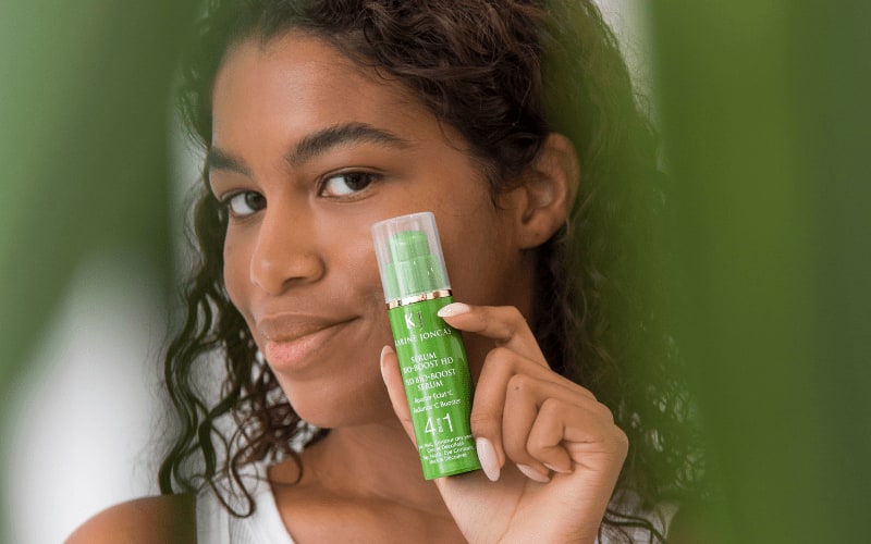 Woman holding a green bottle of Karine Joncas Bio-Boost HD serum next to her cheek