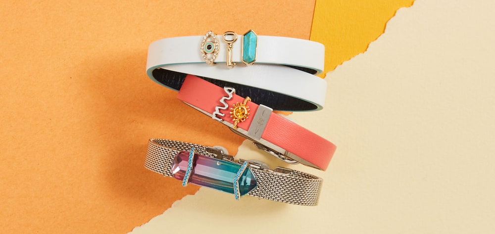Three KEEP bracelets, in white leather, coral leather and silver mesh, decorated with slide-on charms on an orange and cream paper background