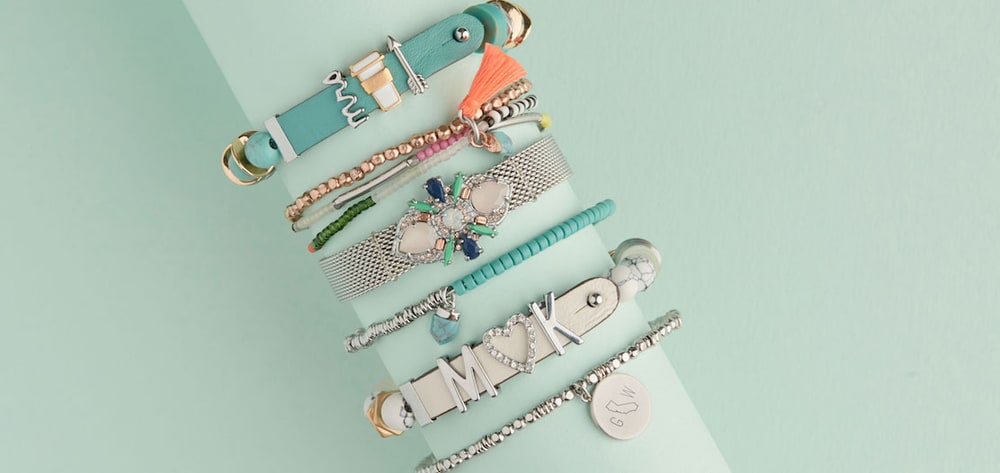 A stack of KEEP bracelets in mint, silver and turquoise, including a mesh band with a jeweled charm and a leather band spelling initials around a heart