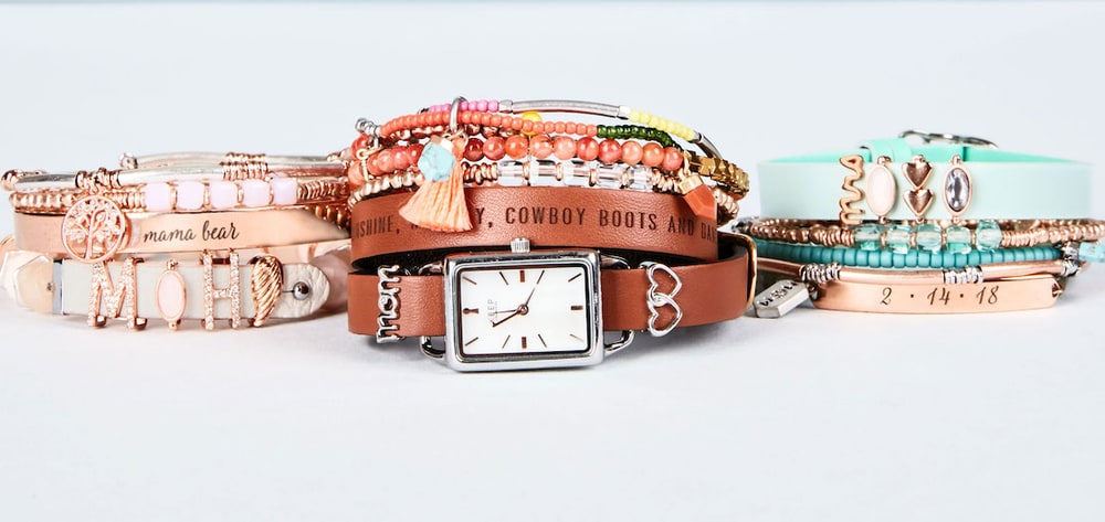 Three stacks of KEEP bracelets and a leather watch band with a mom charm, lined up on a pale grey surface