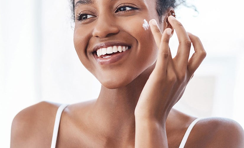 Smiling woman dabbing moisturizer onto her cheek in a bright room