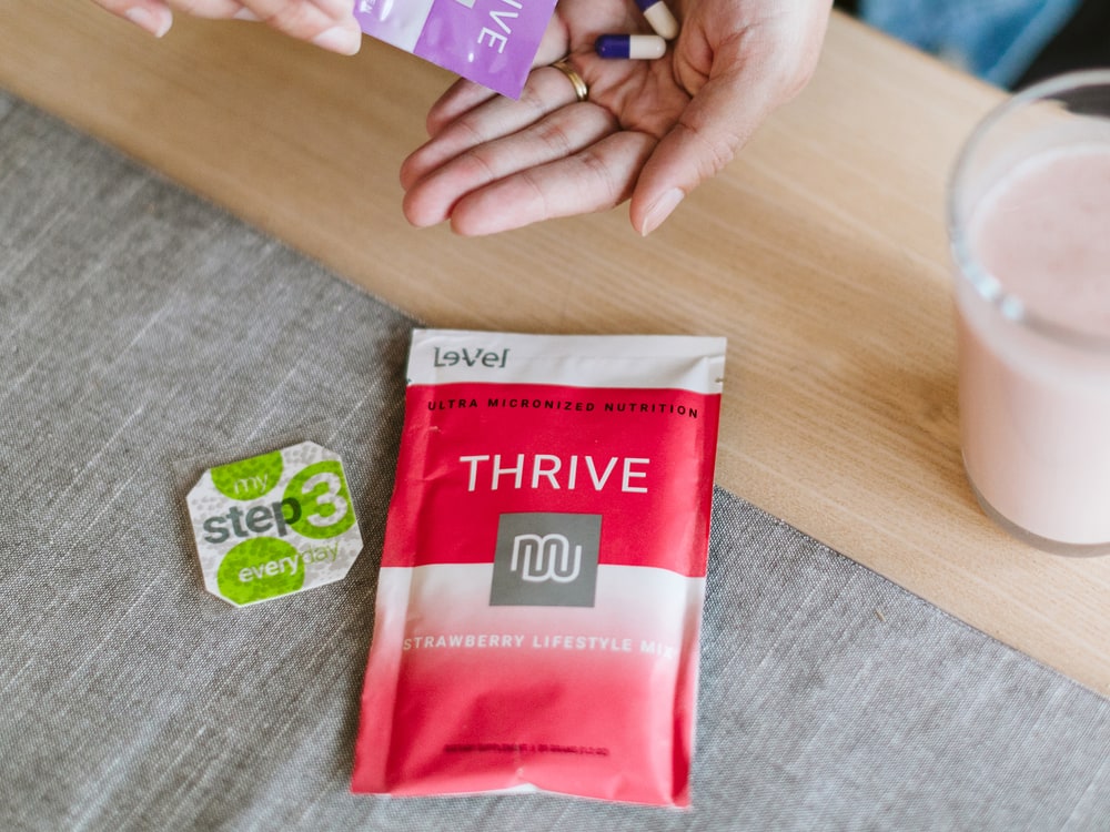 Hands holding the three steps of the Classic THRIVE Experience: a Premium Lifestyle Capsule packet, a Lifestyle Mix shake and a DFT patch