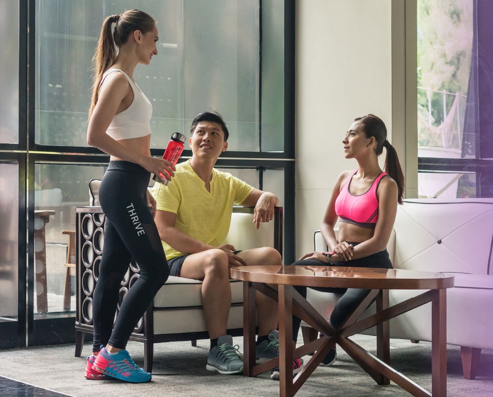Three people in workout clothes talking in a gym lounge, one holding a THRIVE water bottle