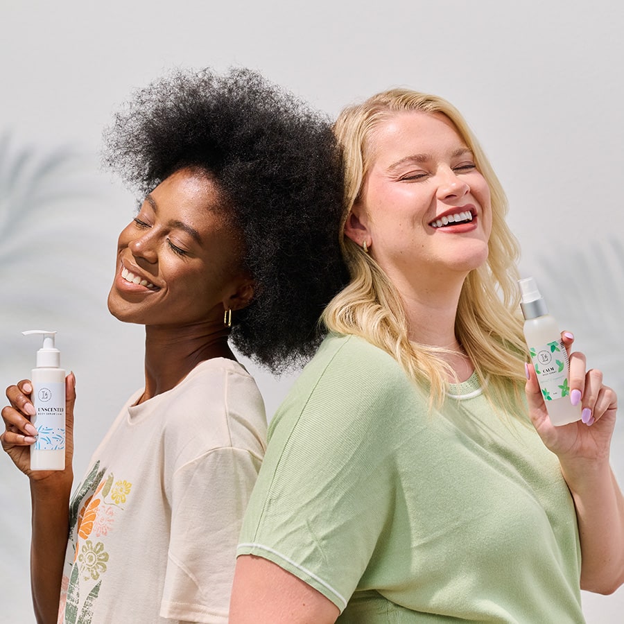 Two laughing women standing back to back, each holding a Lemongrass Spa bottle