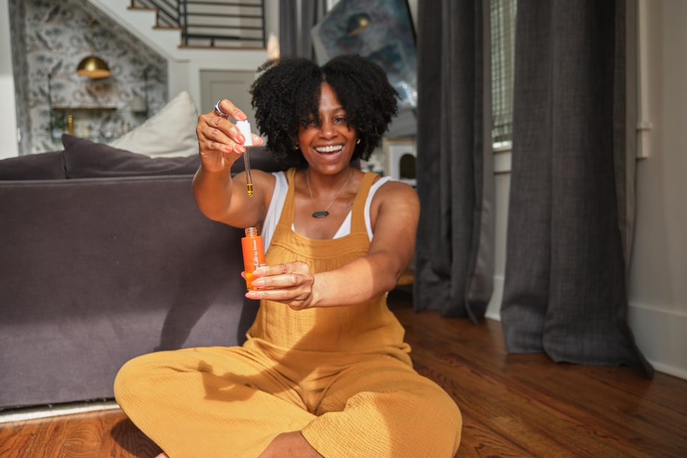 A smiling woman in yellow overalls sits on a living-room floor holding up a LimeLife facial oil and its dropper
