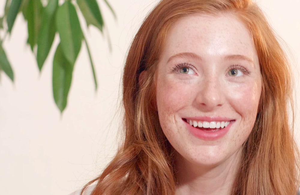 A smiling woman with long red hair and freckles in front of a pale pink wall beside a green plant