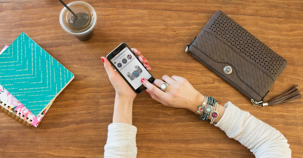 A woman's hands holding a phone over a wooden table, wearing snap bracelets and rings next to a wallet and notebook