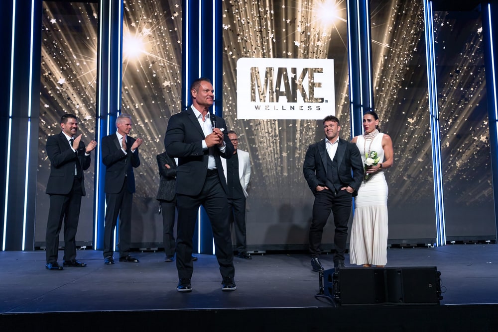A MAKE Wellness leader speaking on stage in front of the company logo at a MAKE event, with executives applauding behind him