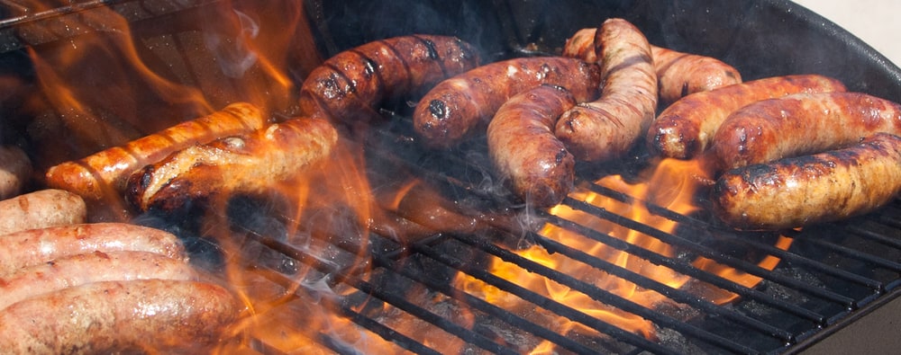 Bratwursts sizzling over open flames on a round charcoal grill
