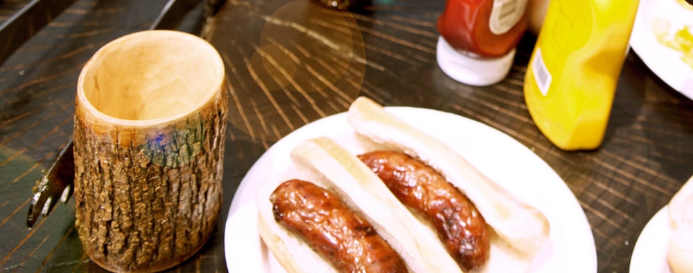 Two grilled brats in buns on a white plate next to a wooden mug made from a tree branch