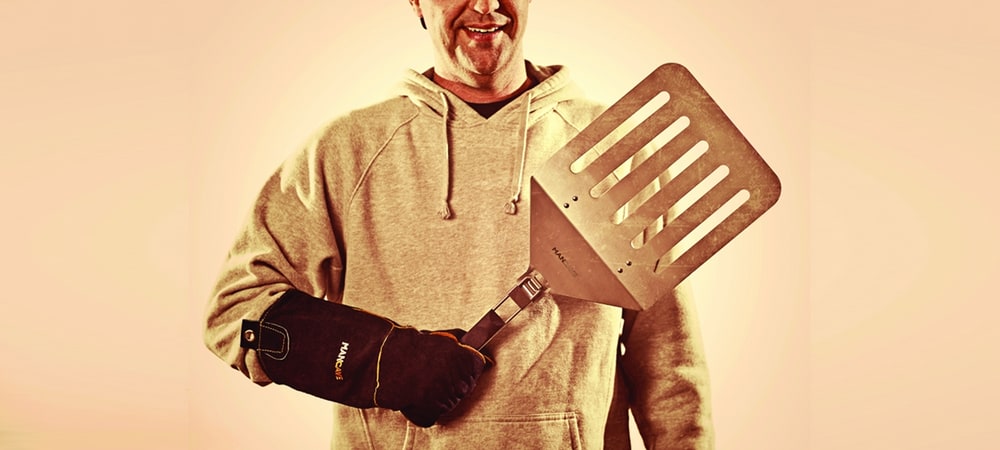 A smiling man in a grey hoodie and leather grill glove holding an oversized stainless steel spatula