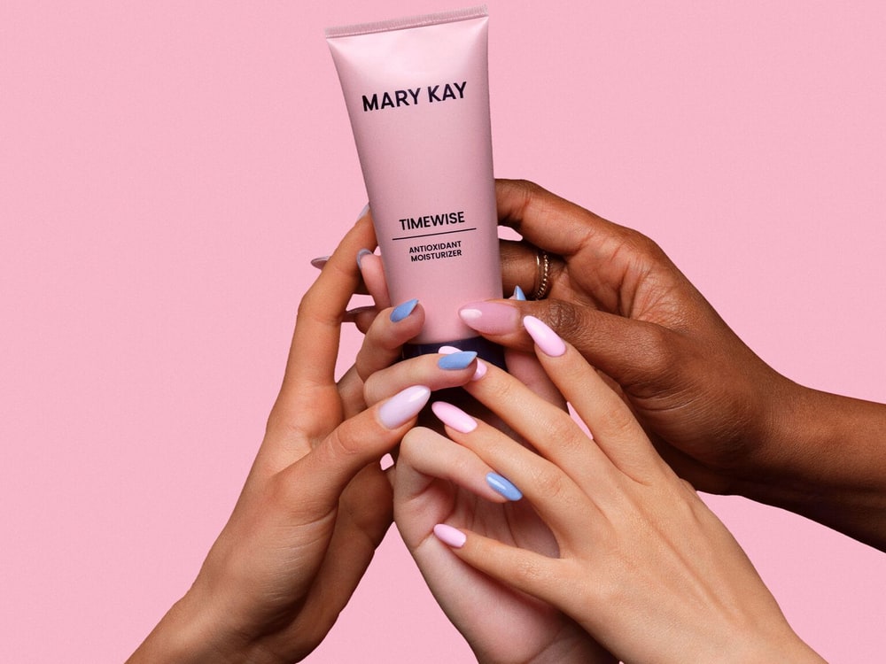 Several hands with pastel nails holding up a tube of Mary Kay TimeWise Antioxidant Moisturizer against a pink background
