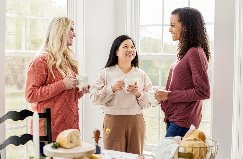 Three smiling women in autumn sweaters chatting over mugs of coffee in a bright room with tall windows