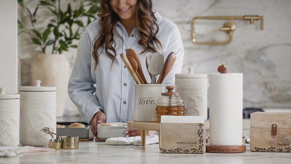 A smiling woman at a marble kitchen counter arranging cream ceramic canisters, a utensil crock of wooden spoons and a paper towel holder
