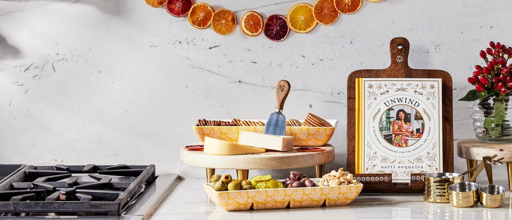 A charcuterie spread on a scalloped yellow serving tray and a two-tier wooden stand on a marble kitchen counter beside brass measuring cups