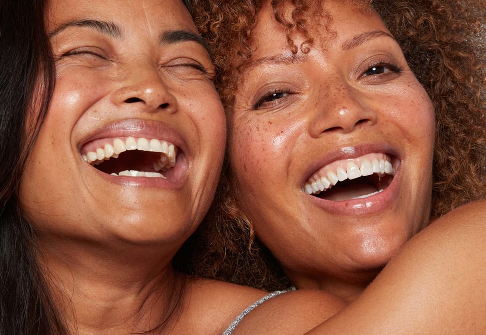 Two smiling women with glowing skin hugging and laughing together