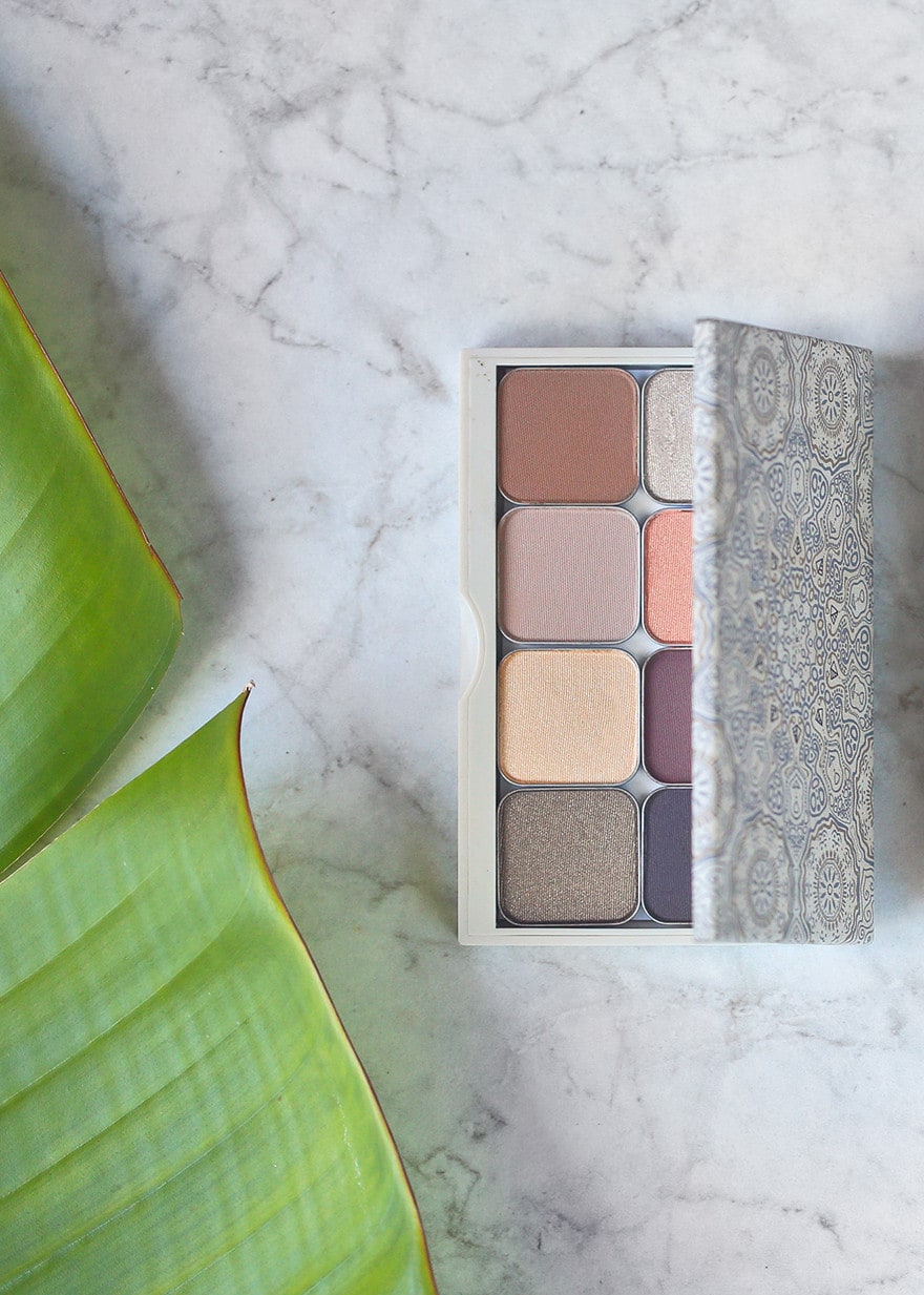 A refillable Maskcara palette of eight eyeshadows open on a white marble counter beside a green leaf