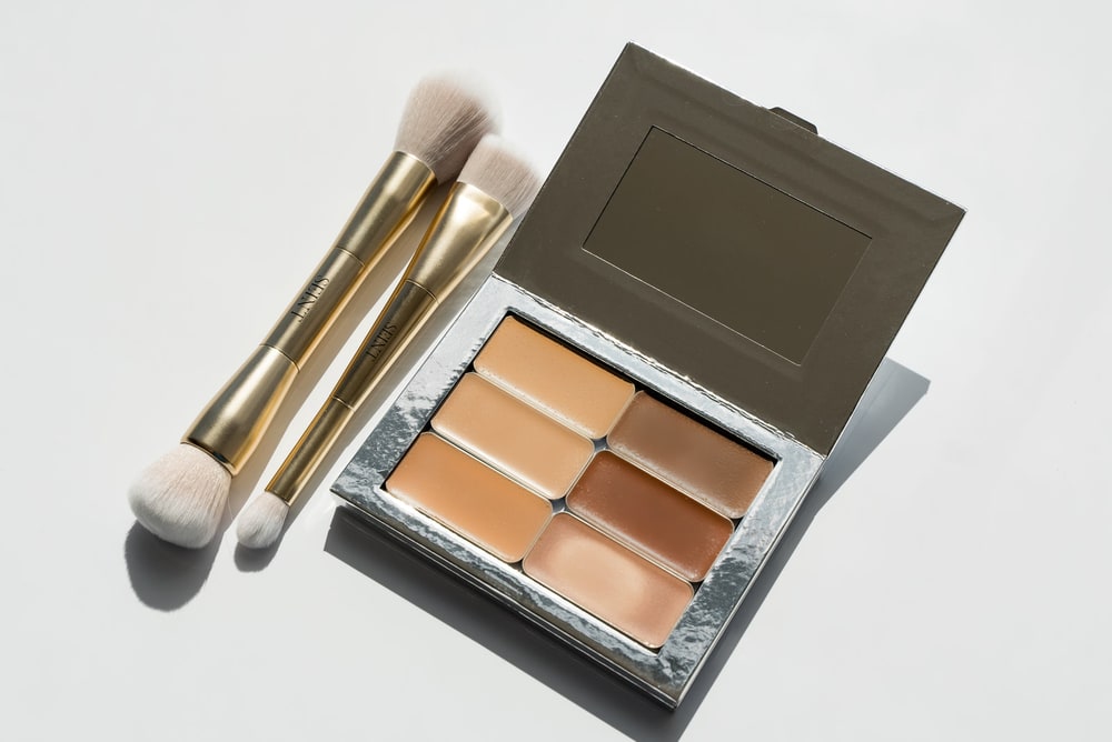 An open Seint palette of six cream shades next to two gold double-ended makeup brushes