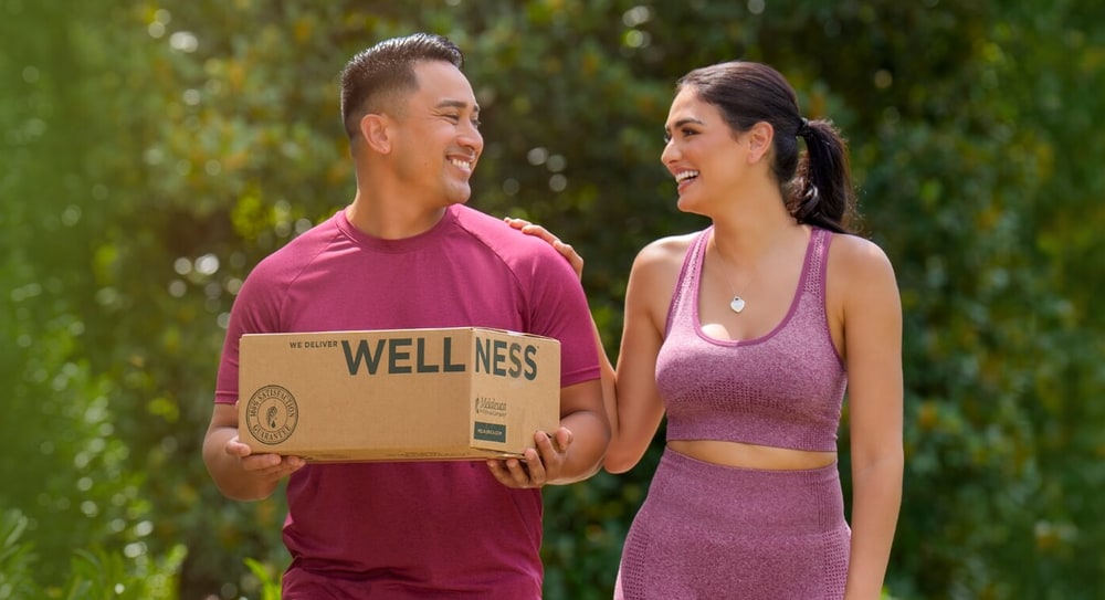 Smiling couple outdoors, the man carrying a Melaleuca delivery box