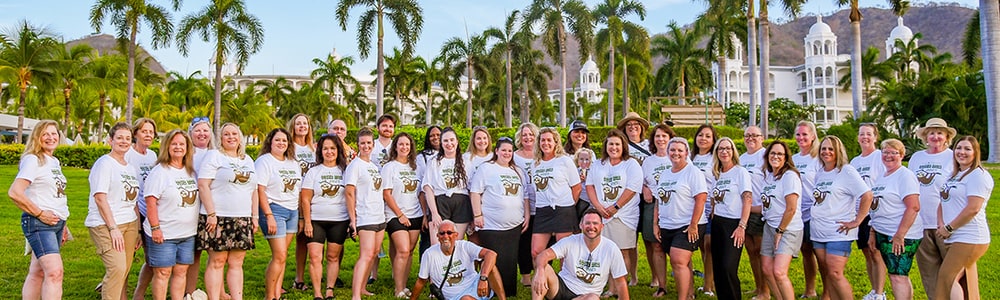 A large group of Mia Bella's Distributors in matching white T-shirts on a lawn lined with palm trees during a company incentive trip to Costa Rica