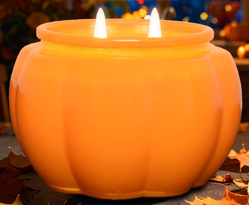 A lit pumpkin-shaped Mia Bella's candle in the Perfect Pumpkin fragrance on a table scattered with autumn leaves