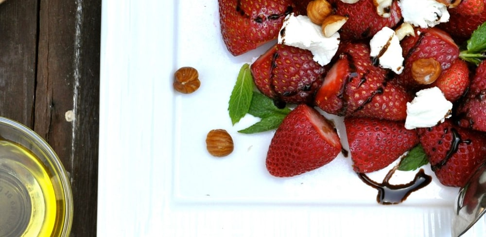 Strawberries with goat cheese, hazelnuts and mint drizzled with dark balsamic on a white platter beside a bowl of olive oil