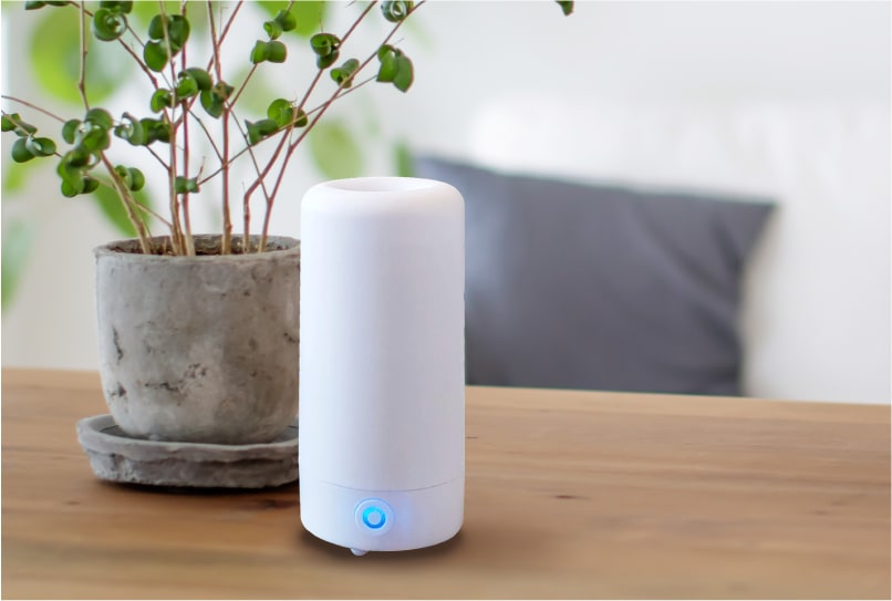 A white AirMoji fragrance diffuser glowing blue on a wooden table next to a potted plant
