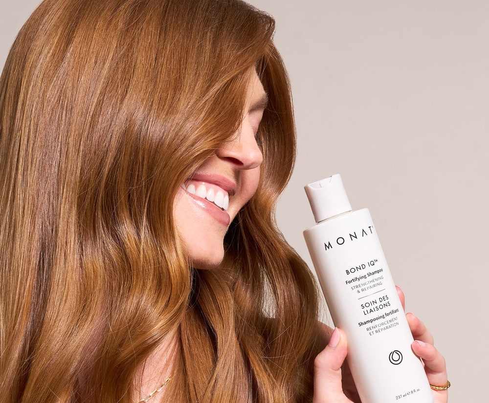 Smiling woman with long auburn hair holding a white bottle of MONAT BOND IQ Fortifying Shampoo