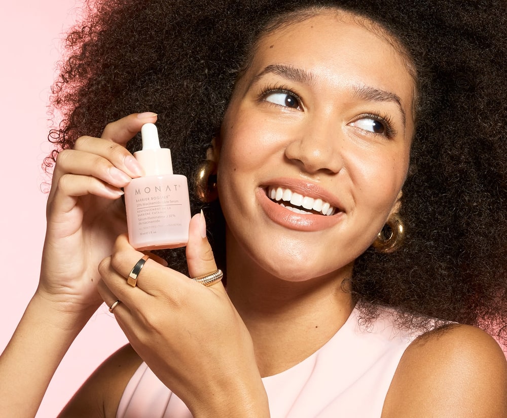 Smiling woman with curly hair holding a pink dropper bottle of MONAT Barrier Booster niacinamide serum