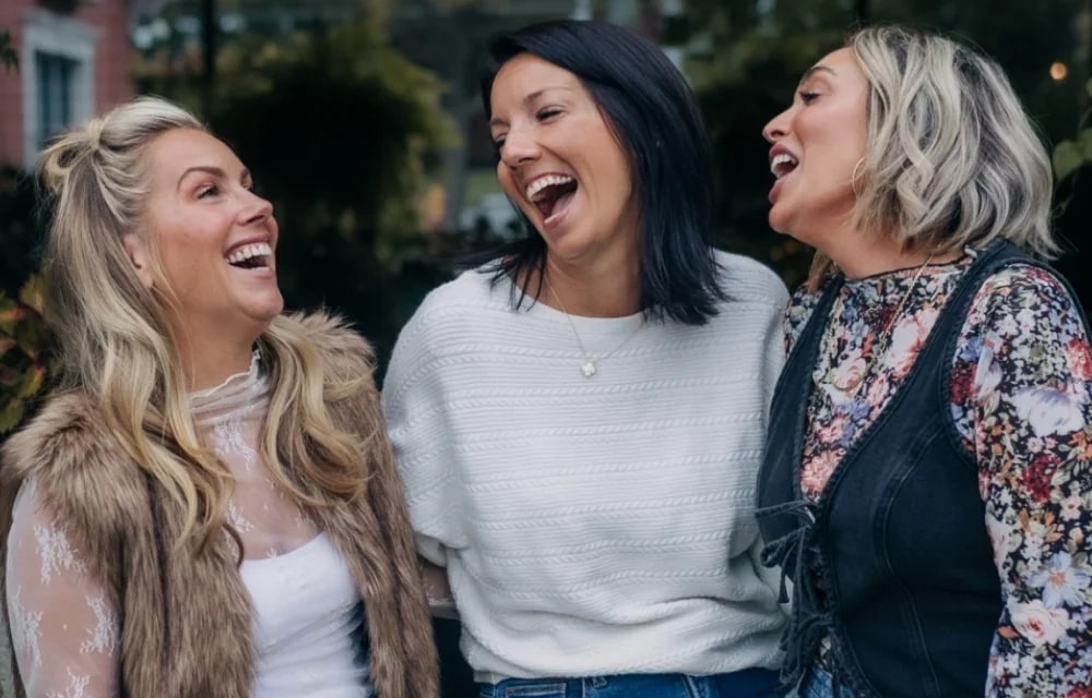 Three smiling women laughing together outdoors in front of trees
