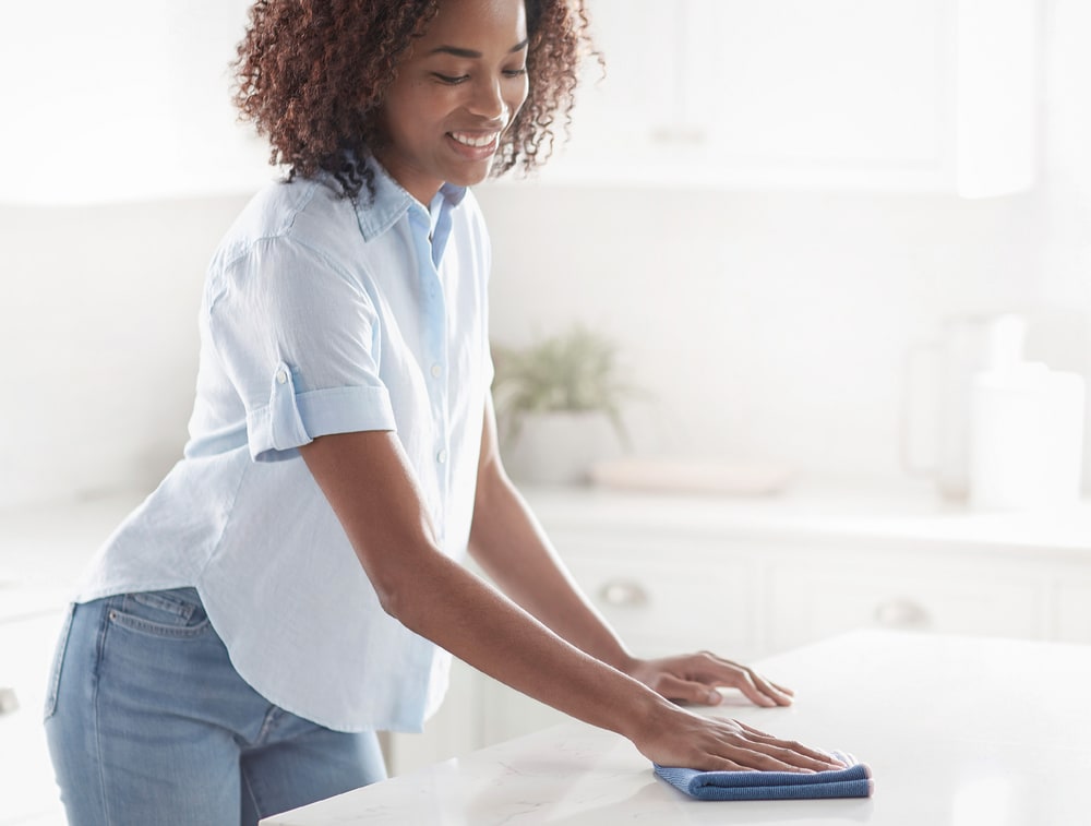 A smiling woman wipes a white kitchen countertop with a blue Norwex microfiber cloth