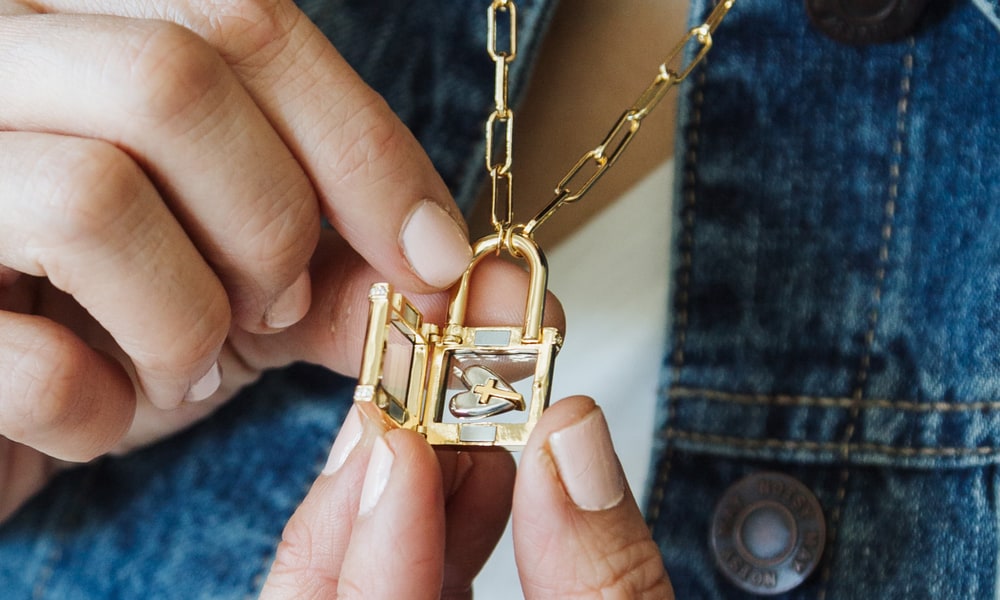 Two hands opening a gold padlock-shaped Origami Owl locket with a small charm inside, worn over a denim jacket