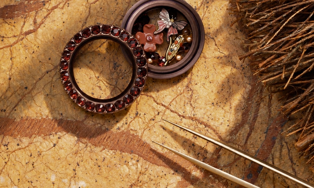 An open bronze Origami Owl Living Locket with red crystals, holding a butterfly, flower and feather charm on a marble surface