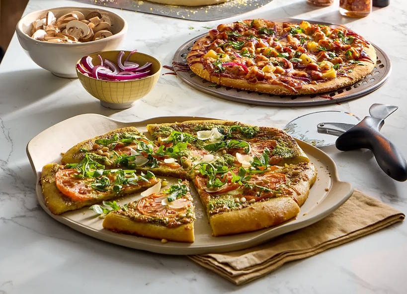 Two pizzas on Pampered Chef stoneware pizza stones with a pizza cutter and prep bowls