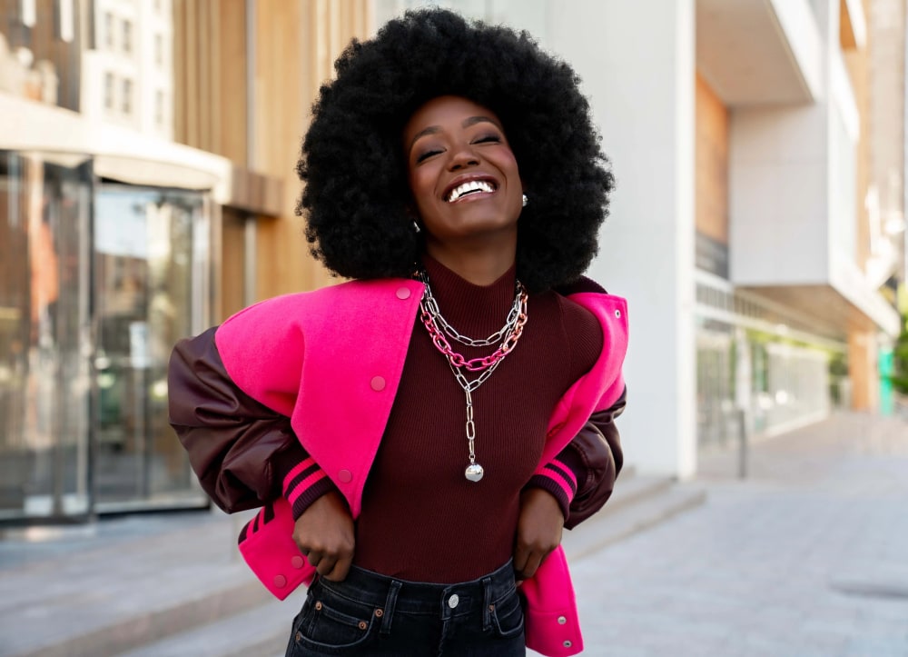 Smiling woman in a pink jacket wearing layered Paparazzi chain necklaces on a city street