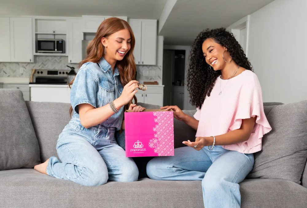 Two women on a couch opening a pink Paparazzi Accessories starter kit box